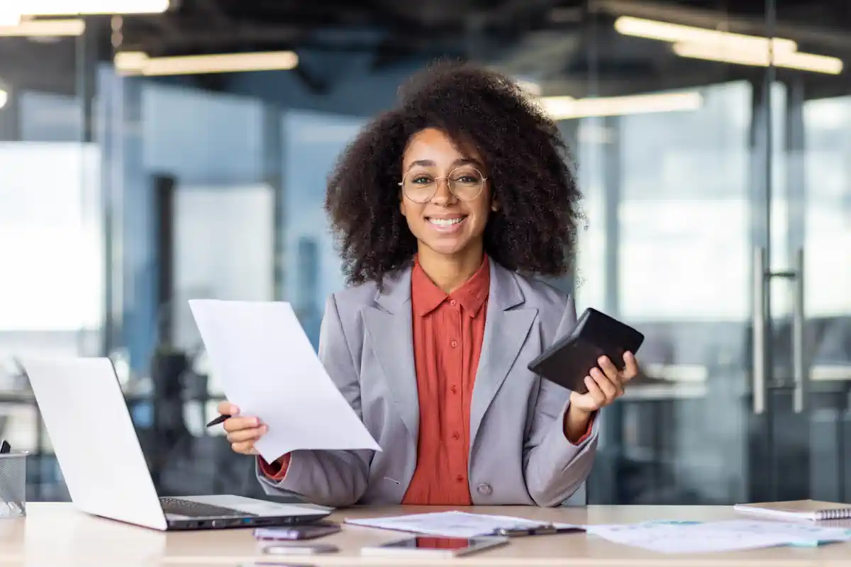 Une femme qui tient une feuille dans une main et une calculatrice dans l'autre assise devant son ordinateur