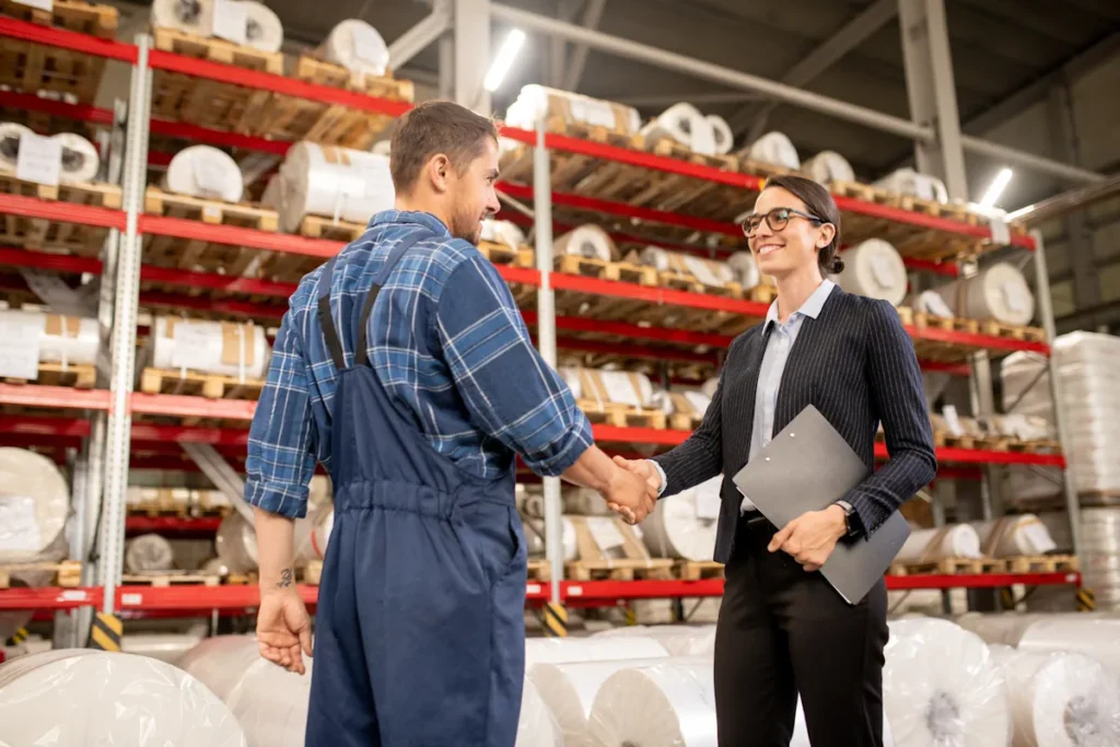 Une femme en costume noir qui serre la main d'un homme en combinaison bleu dans un entrepot
