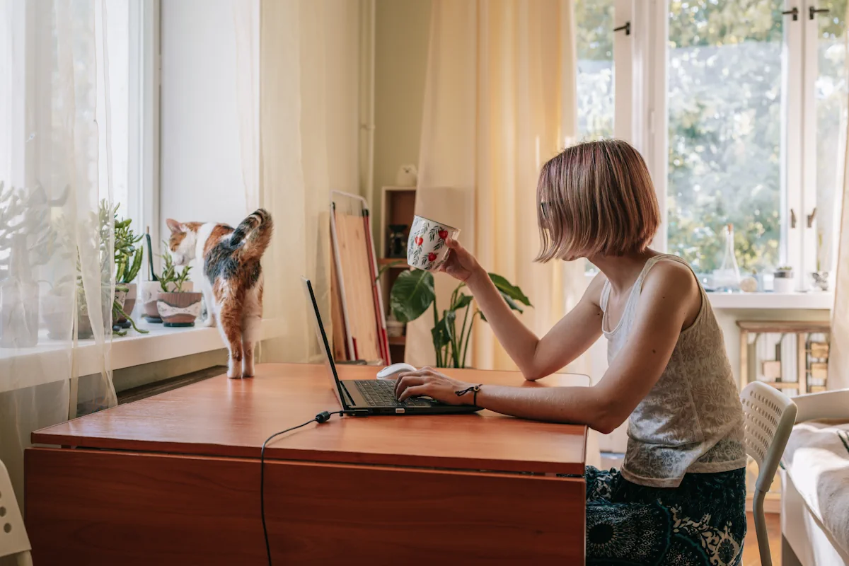 Une femme qui travaille sur son ordinateur en tenant tune tasse dans sa main et son chat devant elle