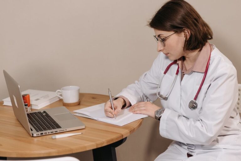 Woman doctor with stethoscope writing at desk with laptop and notes.