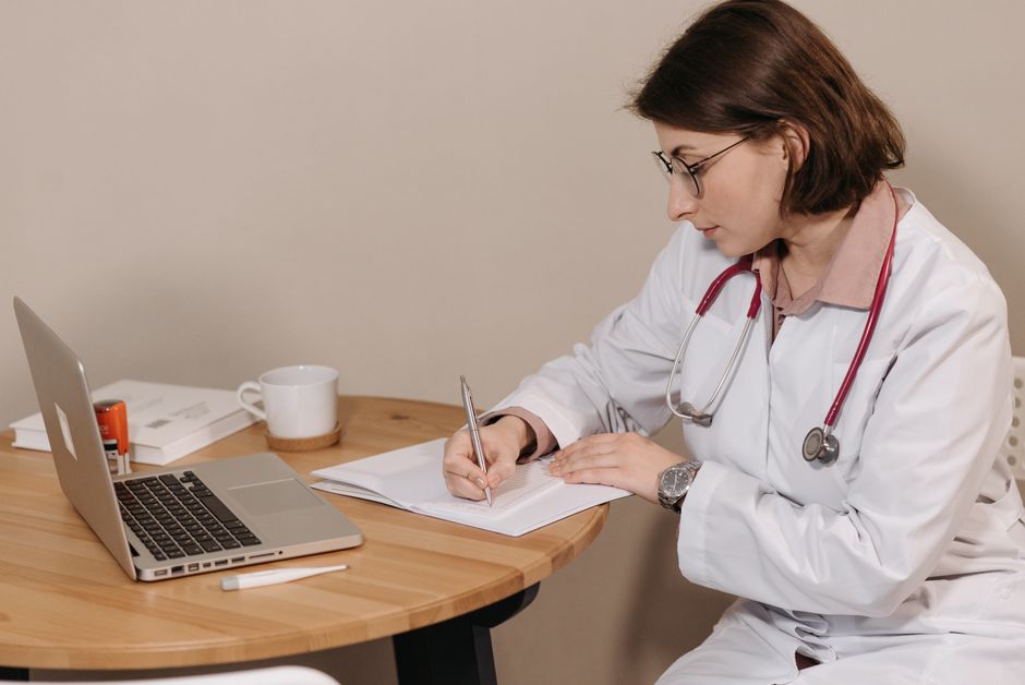 Woman doctor with stethoscope writing at desk with laptop and notes.